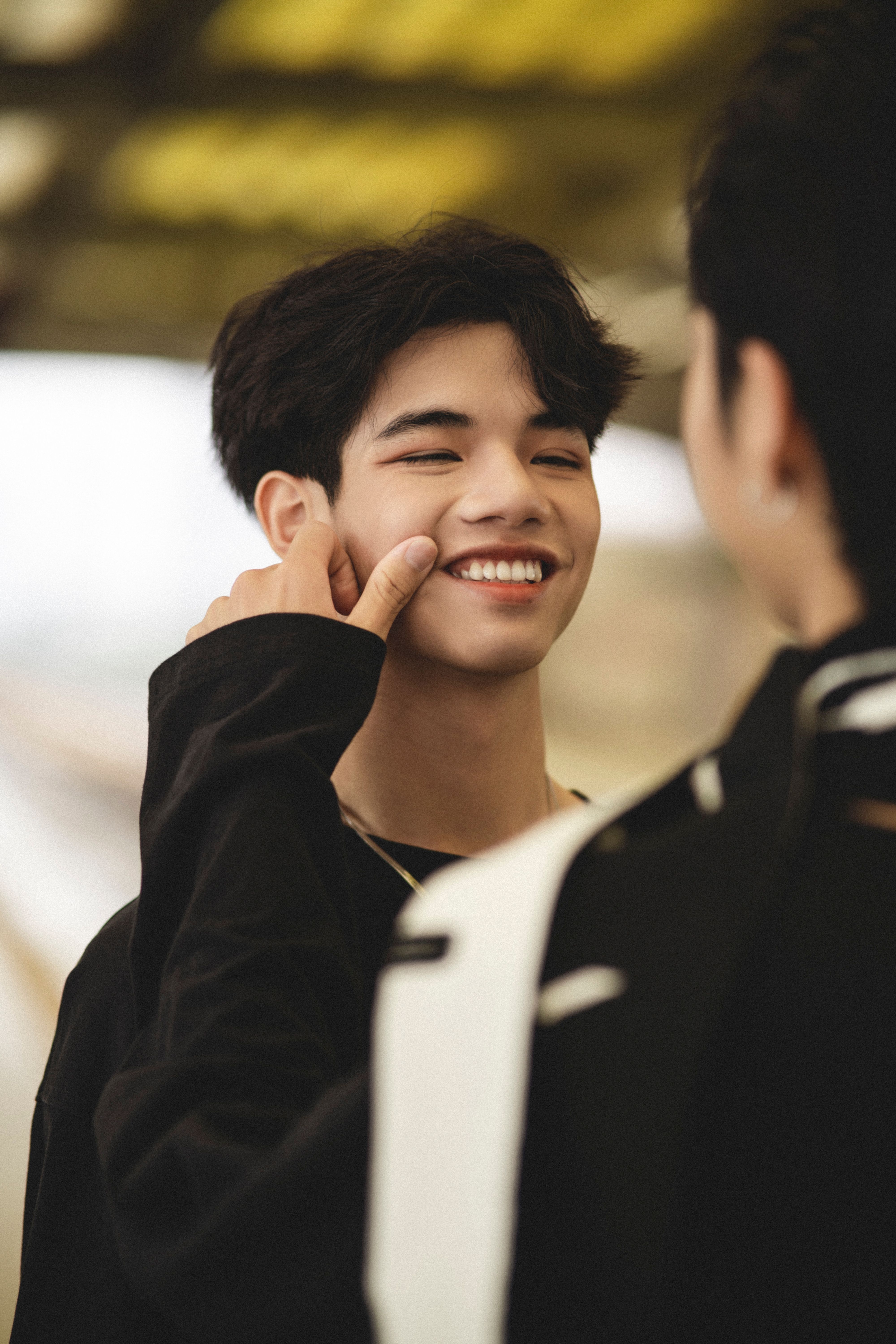 A young man with black hair smiles joyfully as someone gently touches his face in an outdoor setting.