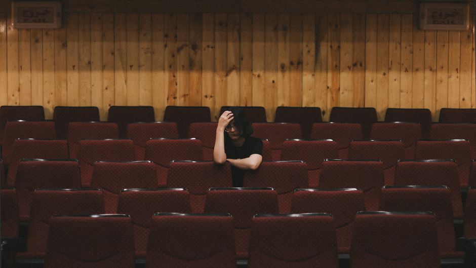 A person sitting alone in a cinema hall with red seats, expressing contemplation.