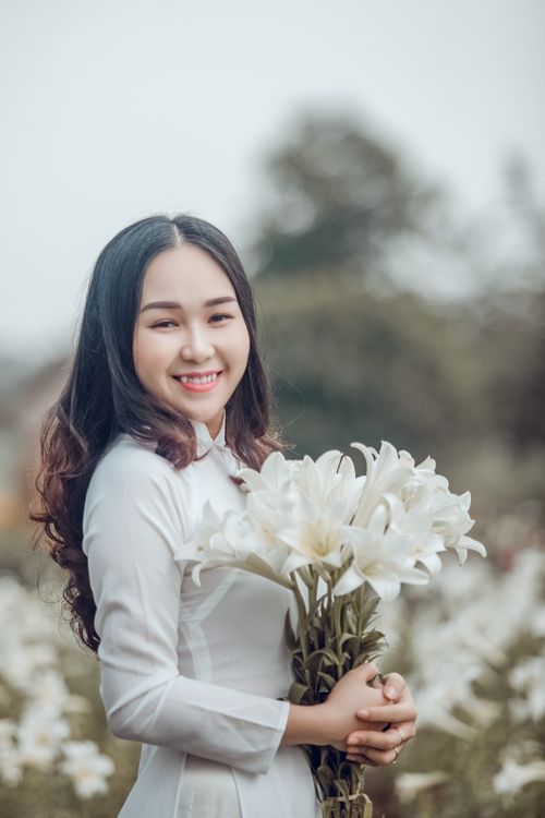Free A cheerful woman in a white dress stands outdoors, holding a bouquet of white lilies and smiling warmly. Stock Photo
