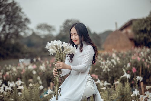 Photo of Woman In a White Outfit Kneeling While Holding White Petaled Flowers