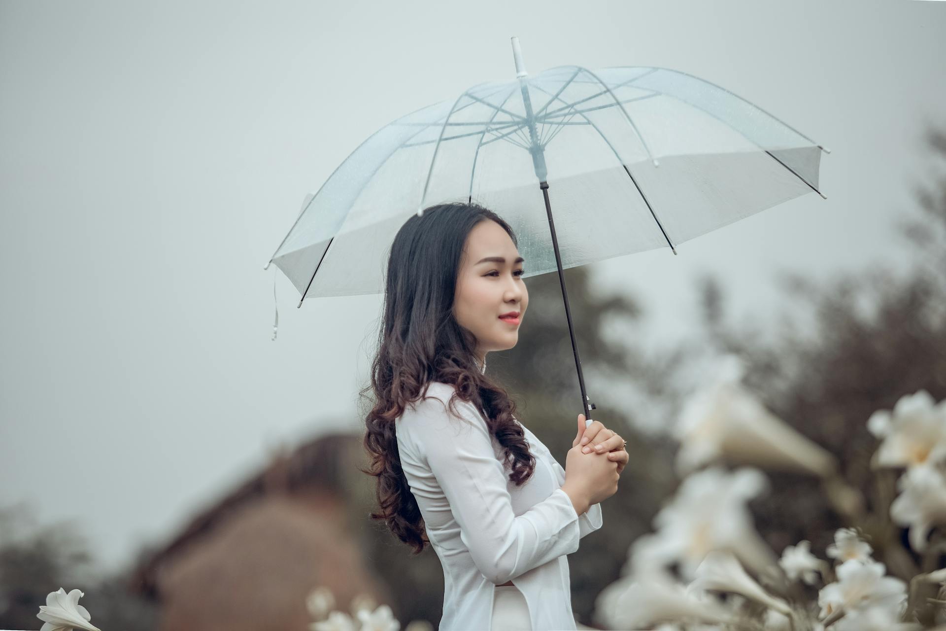 Beautiful Asian woman in white dress posing with umbrella in a serene outdoor setting.