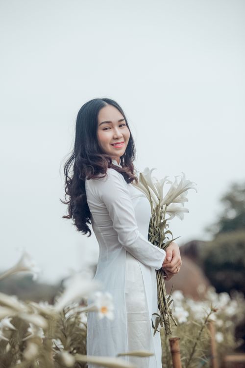 Free Woman in traditional ao dai dress smiles while holding flowers in a serene outdoor setting. Stock Photo