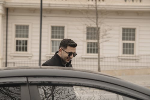 A man in sunglasses and a black coat is standing by a car