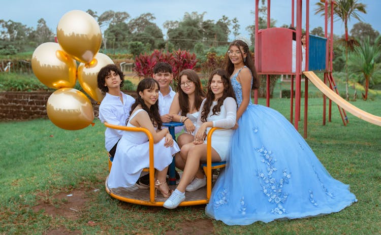 Smiling Women, Girls And Boys Sitting In Shirts And Dresses At Playground