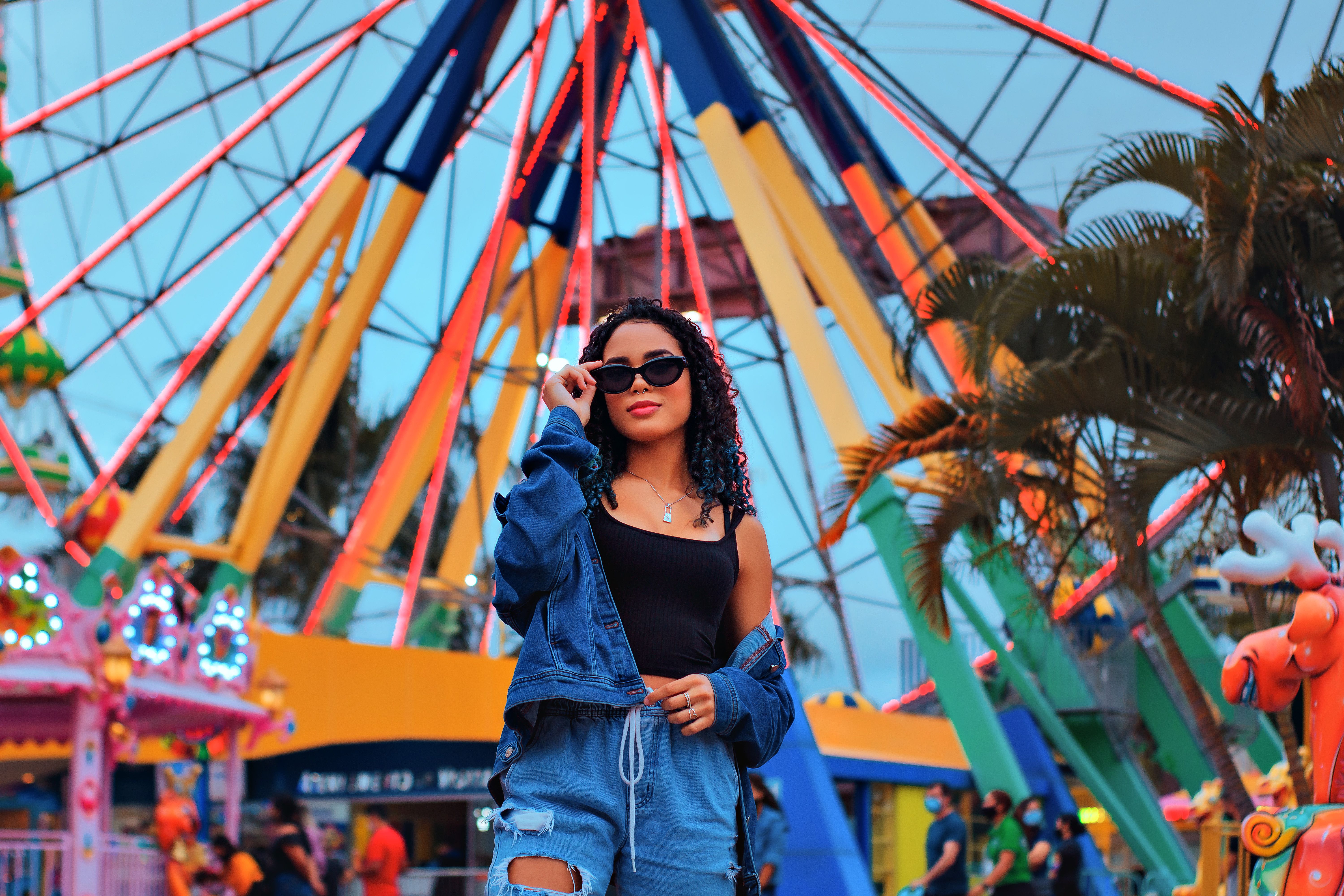 Stylish woman in denim poses at a lively amusement park with colorful Ferris wheel.