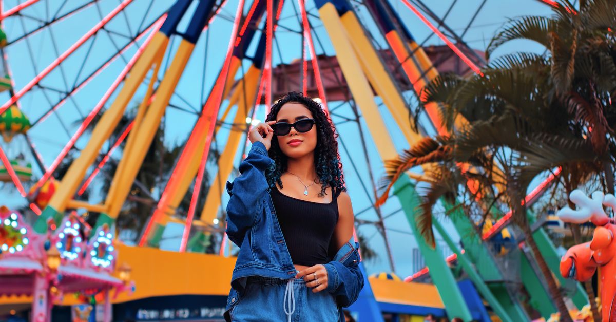 Stylish woman in denim poses at a lively amusement park with colorful Ferris wheel.