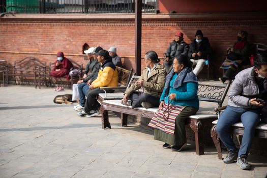 A diverse group of people sitting on benches in a sunlit square in Kathmandu, Nepal.