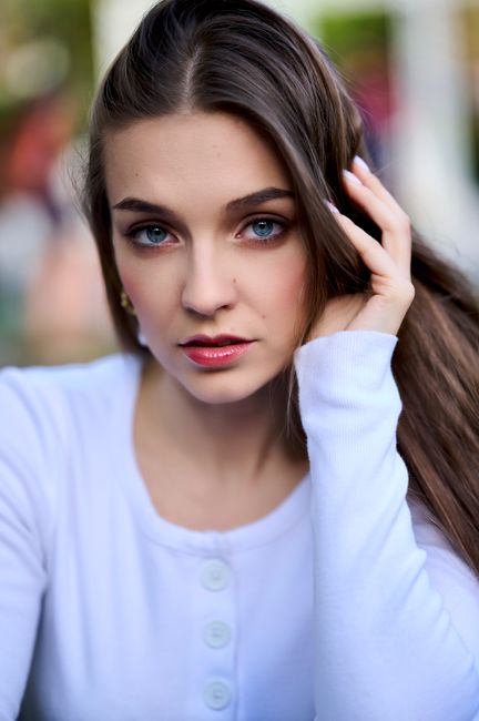 Close-up portrait of a woman with long brown hair and blue eyes wearing a white blouse outdoors.