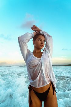 A woman poses on the beach with arms raised, wearing a wet white shirt and bikini bottoms.