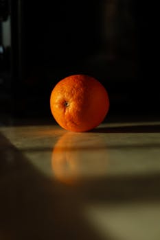 A single orange illuminated in soft light, casting a reflection on a tabletop.