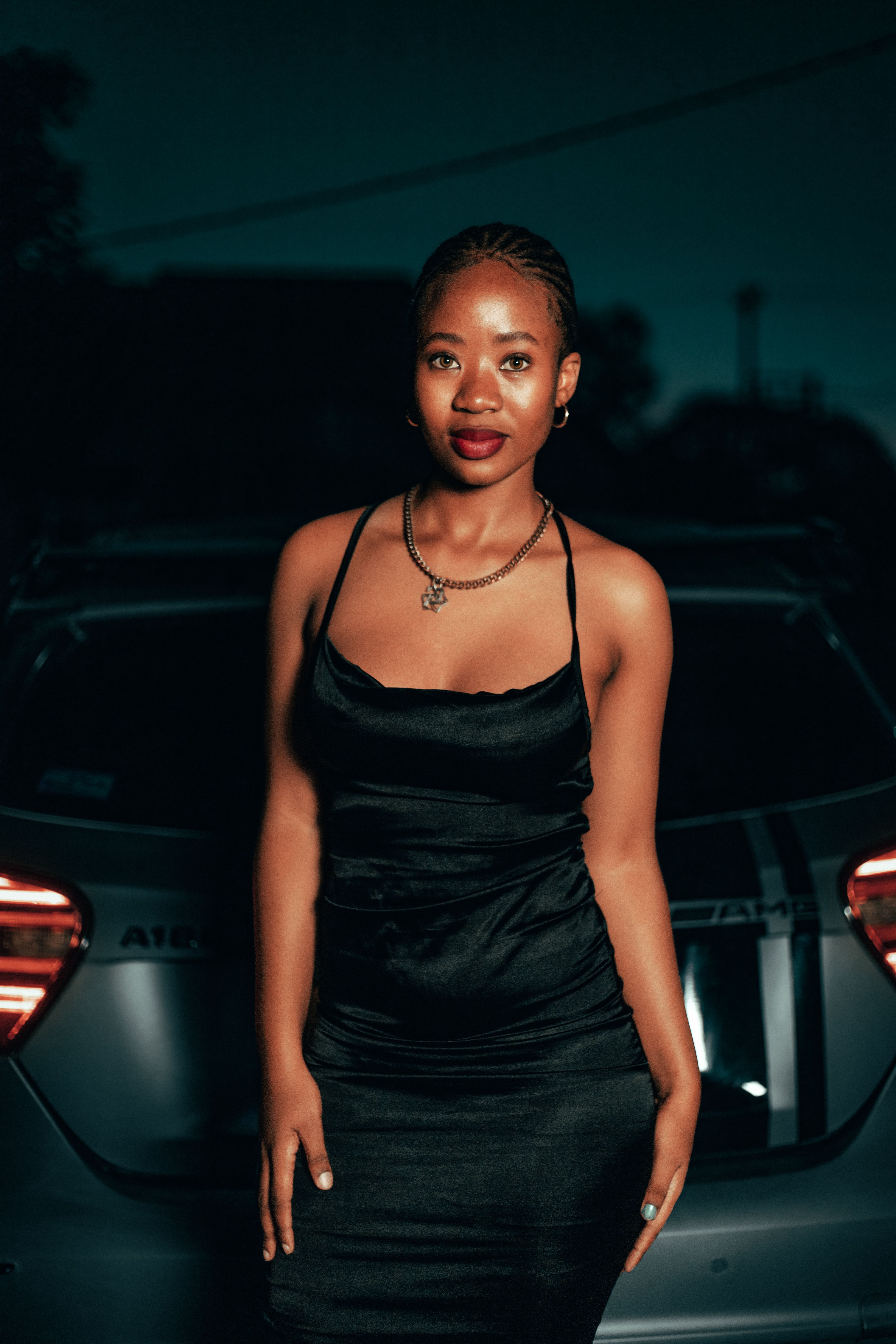 Stylish young woman in a black dress posing confidently against a car backdrop at dusk.