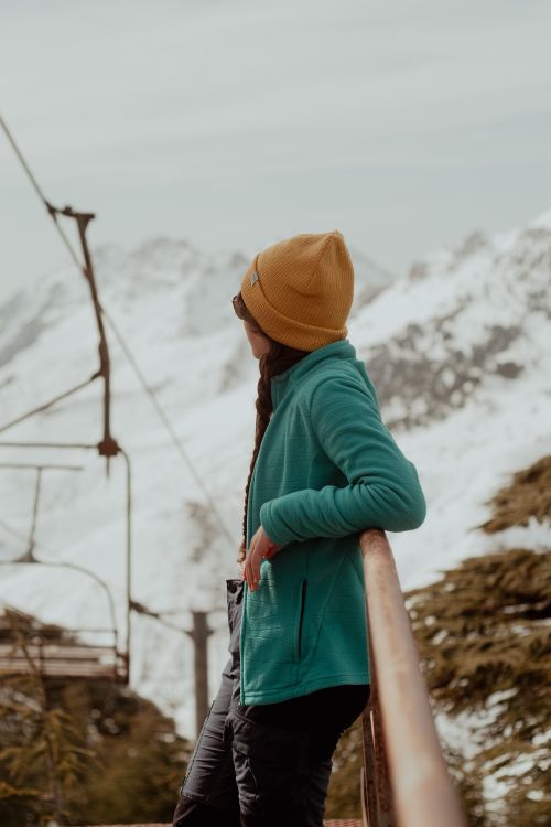 Free A woman in a beanie and jacket stands by a railing, admiring snowy mountains. Stock Photo