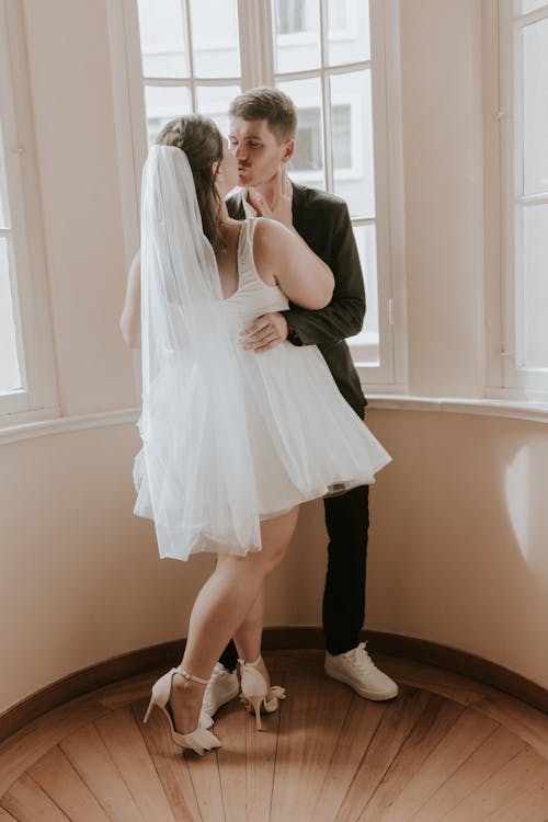 A bride and groom kiss in a room with windows