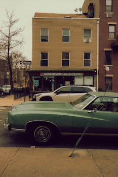 Green vintage car parked on a city street outside a tattoo parlor.
