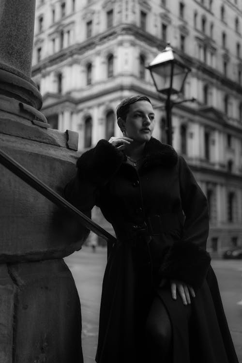 Black and white photo of woman in coat leaning against building