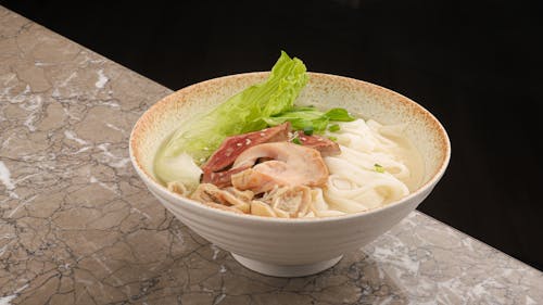 A bowl of noodles with meat and lettuce on a marble counter