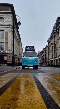 Classic blue Volkswagen minibus parked in a vintage cityscape with cloudy skies.