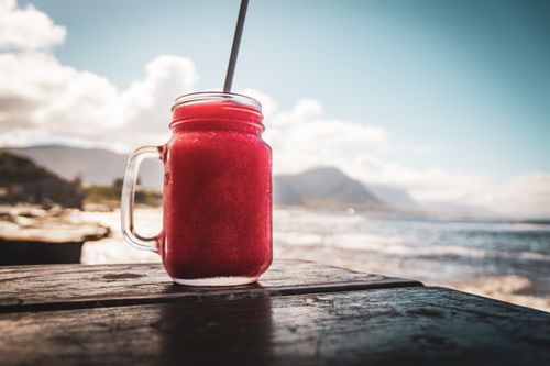 Free A vibrant red berry drink on a wooden table overlooking a scenic coastline on a sunny day. Stock Photo