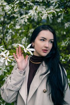 Portrait of a sophisticated woman in a beige coat surrounded by blooming magnolia flowers in spring.