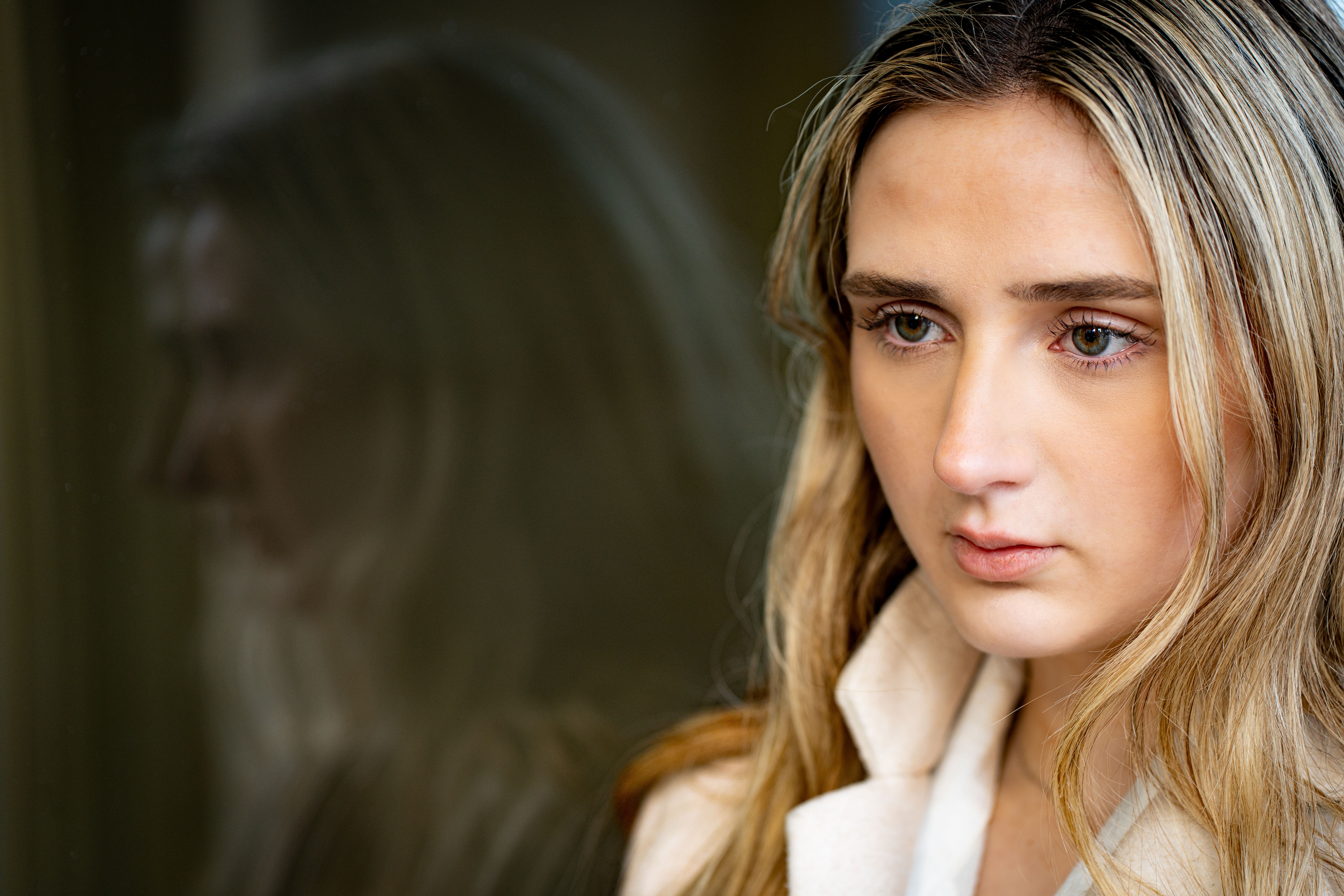 Portrait of a young woman with blonde hair gazing out a window with a reflection.