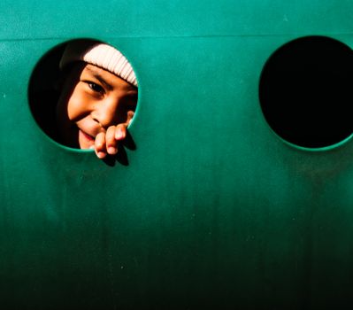 A joyful child peeping through a hole in a playground structure, playfully smiling.