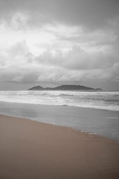 A tranquil beach scene with waves crashing and a distant island under an overcast sky.