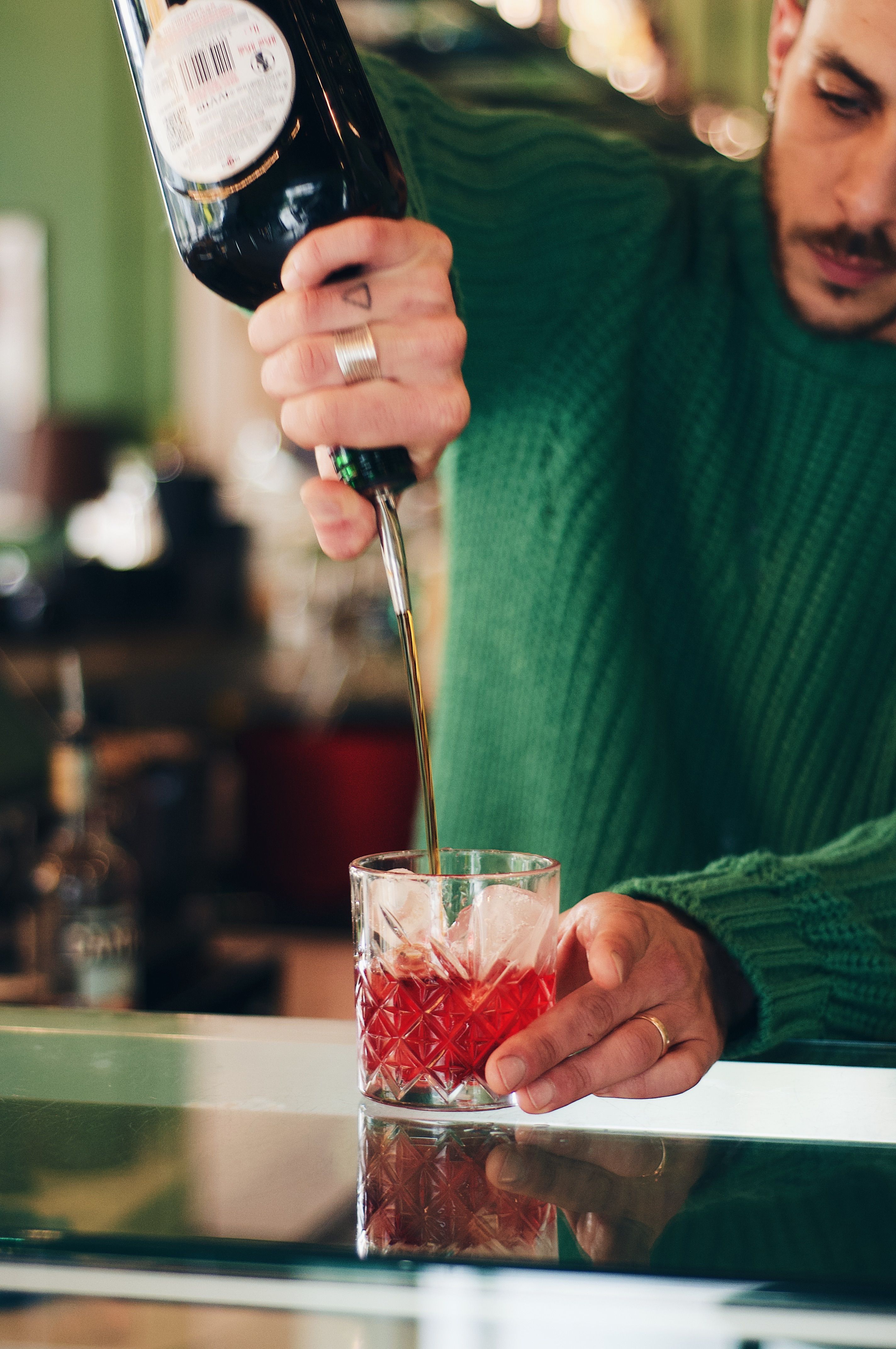 A bartender in a green sweater pours a red cocktail in a glass with ice in a cozy bar.