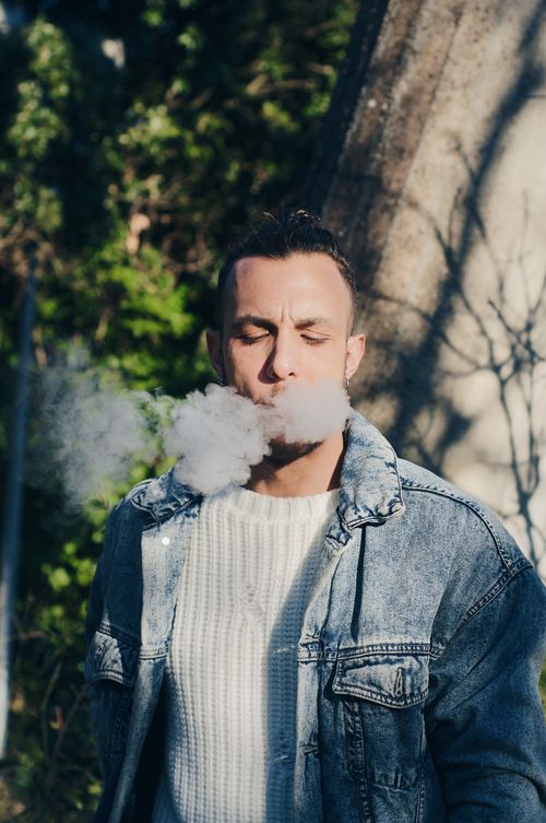 Free A portrait of a young man exhaling smoke outdoors, bathed in sunlight and surrounded by trees. Stock Photo