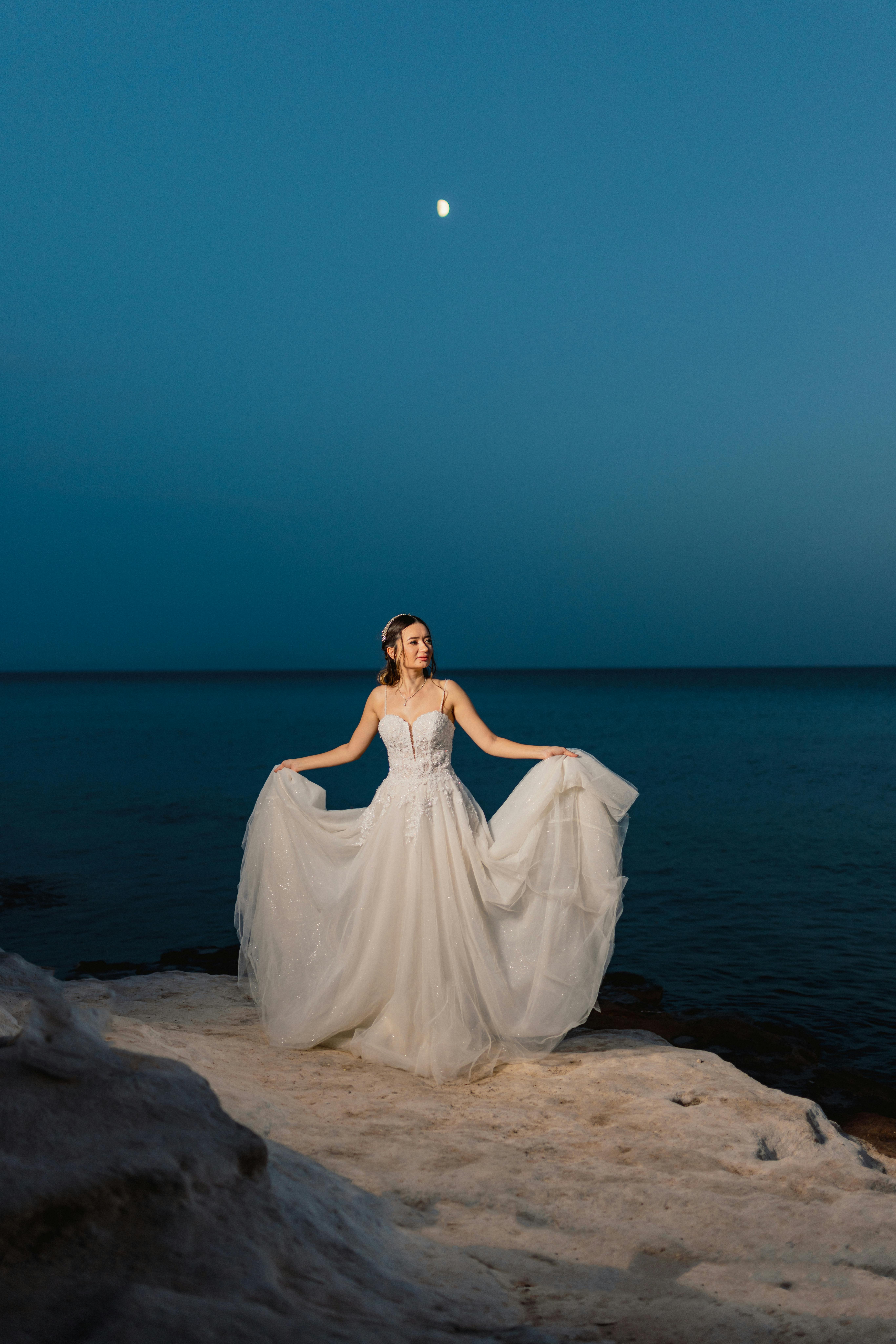 A bride in a gown poses gracefully under a moonlit sky by the beach, evoking elegance and tranquility.