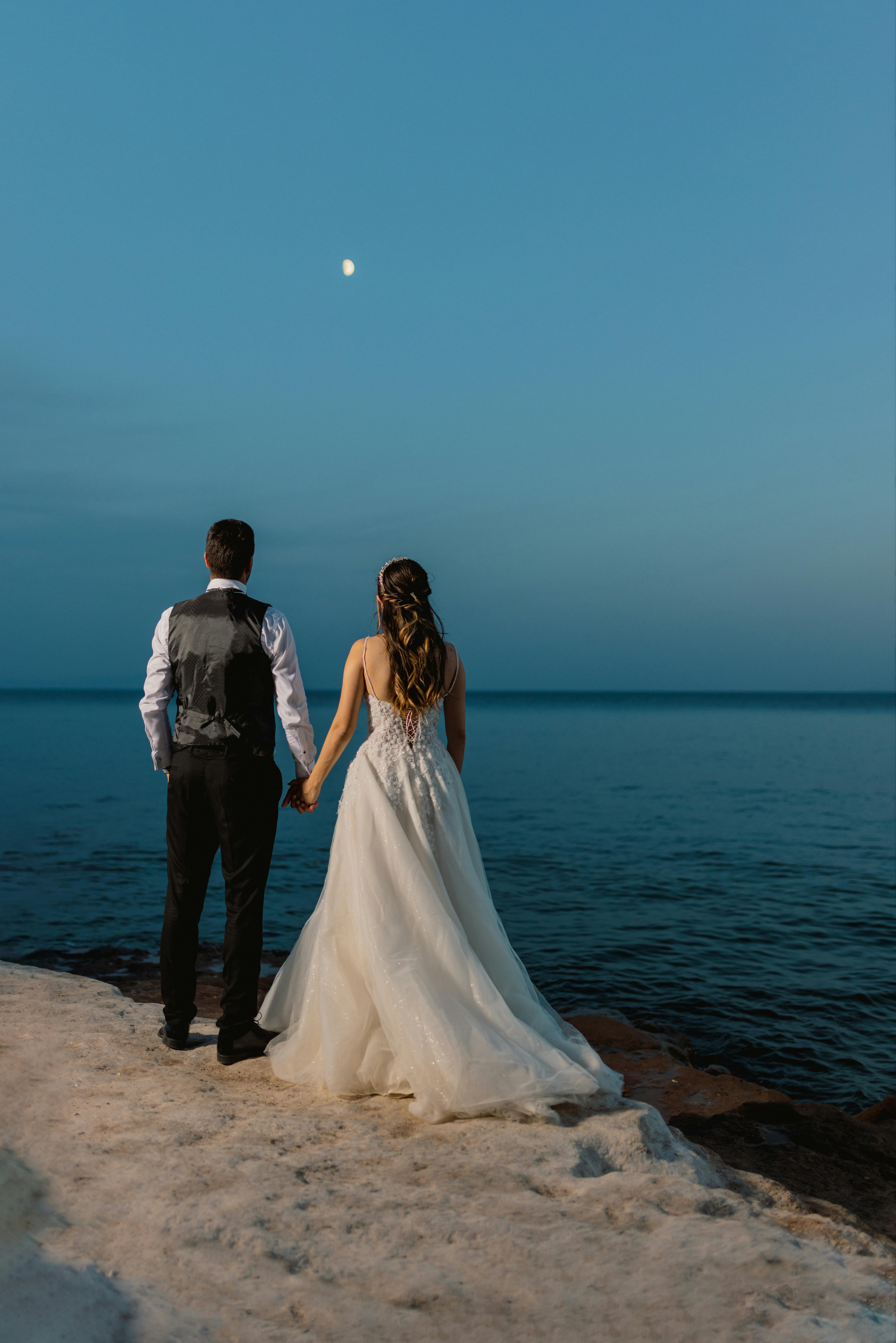 Bride and groom holding hands by the sea under the moonlight, symbolizing love and commitment.
