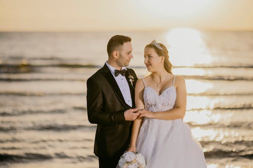 A bride and groom standing on the beach at sunset