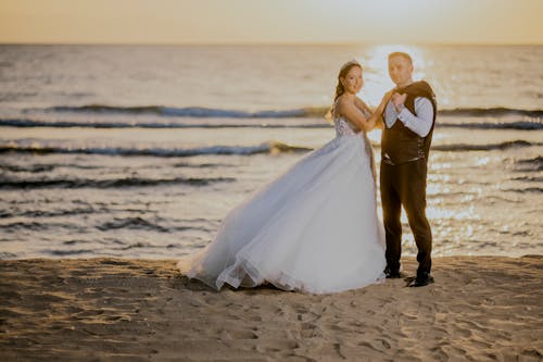 A bride and groom standing on the beach at sunset