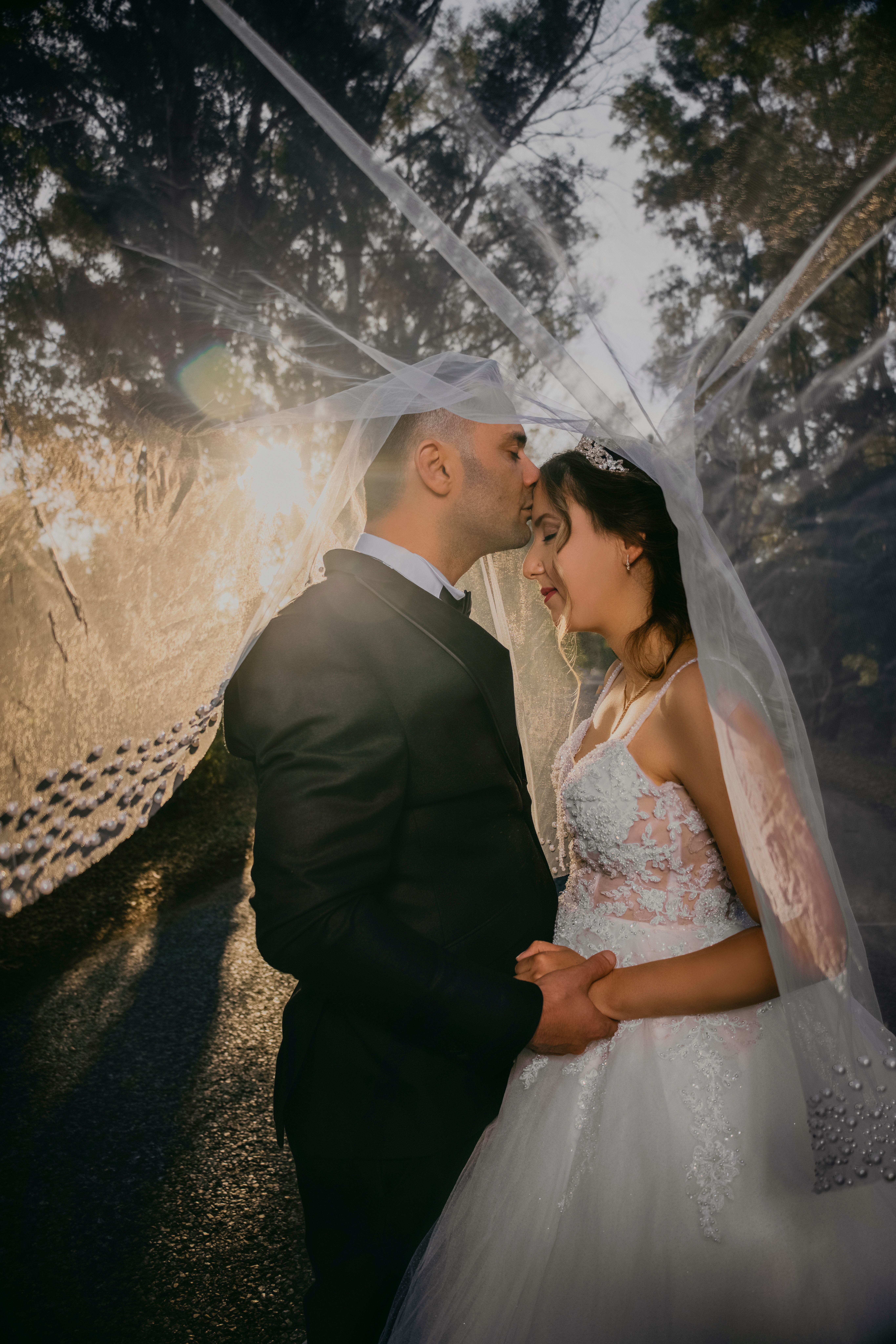 A bride and groom share a tender moment under a veil outdoors during golden hour.