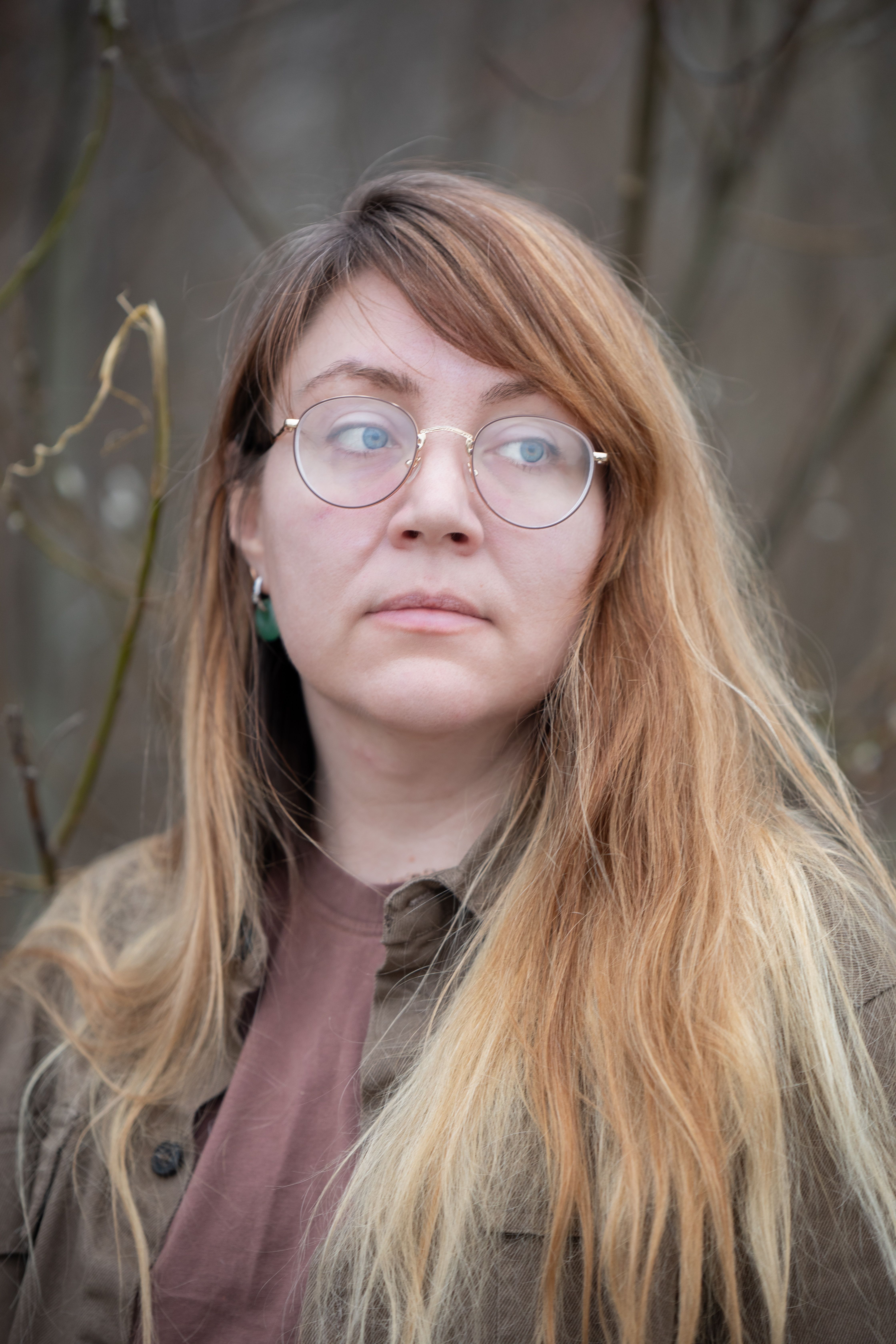 A woman with glasses looking contemplative in a natural outdoor setting.