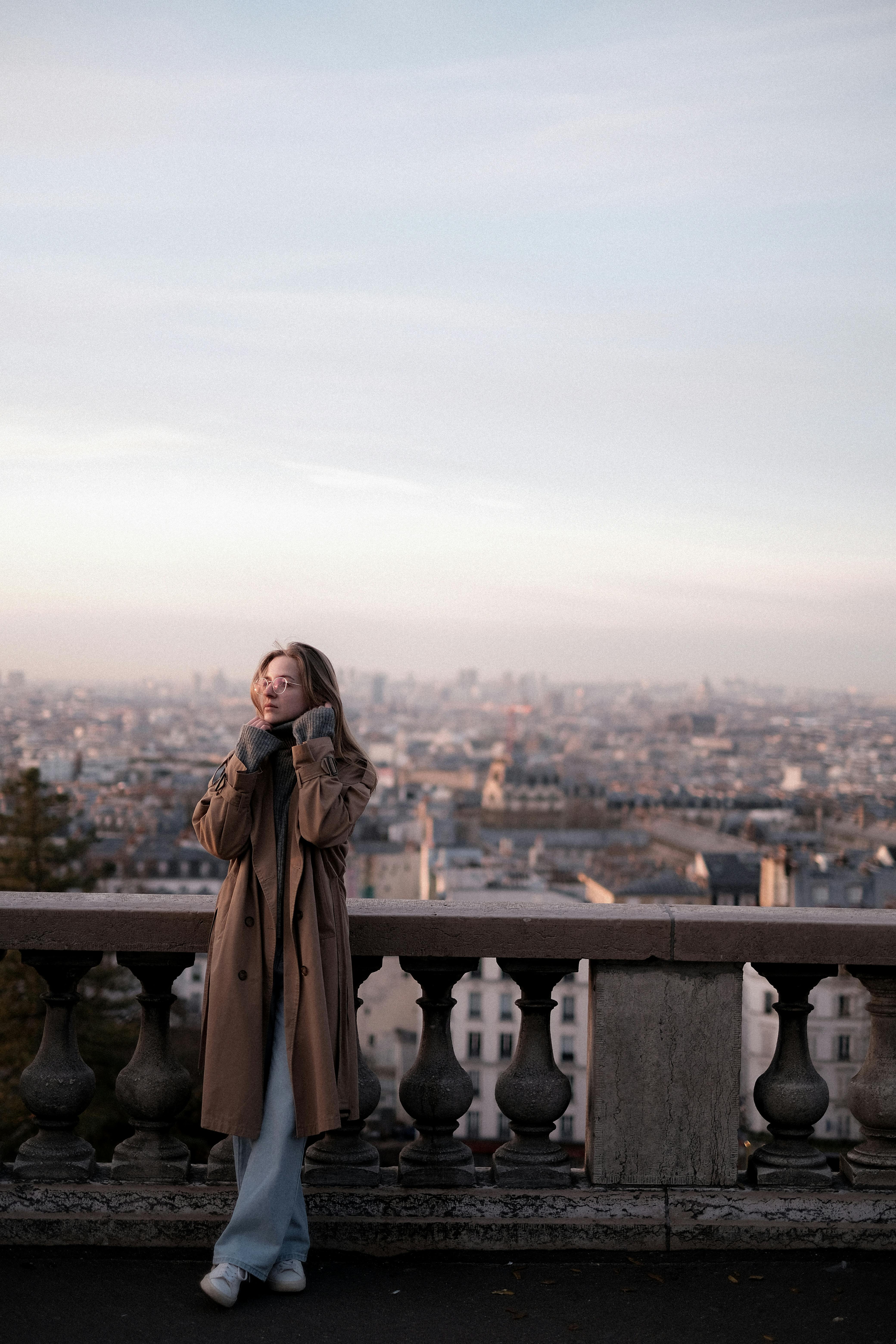 A woman in a trench coat enjoys a scenic city skyline view from a high vantage point during sunset.