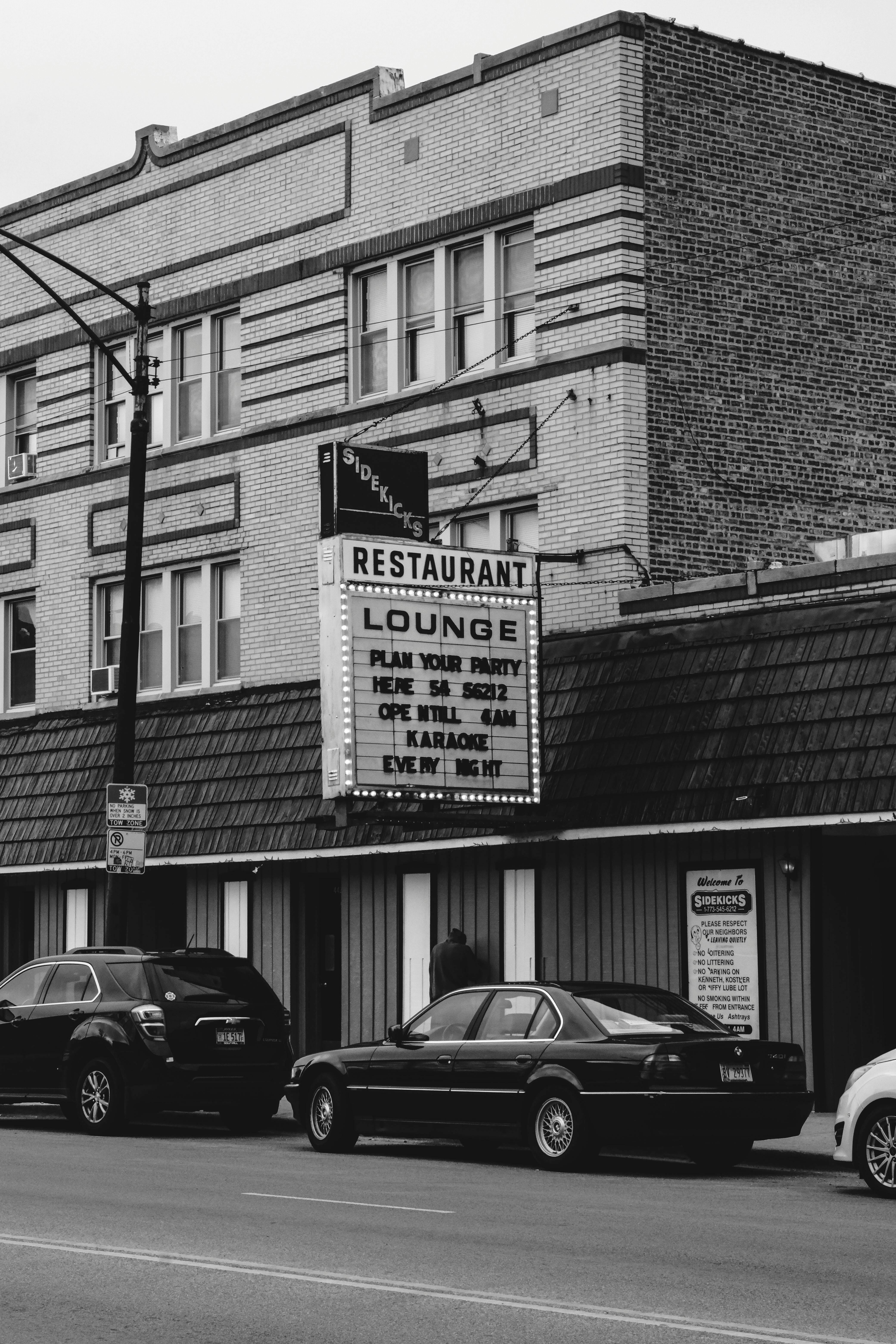 Monochrome view of a vintage restaurant and lounge with cars parked outside.