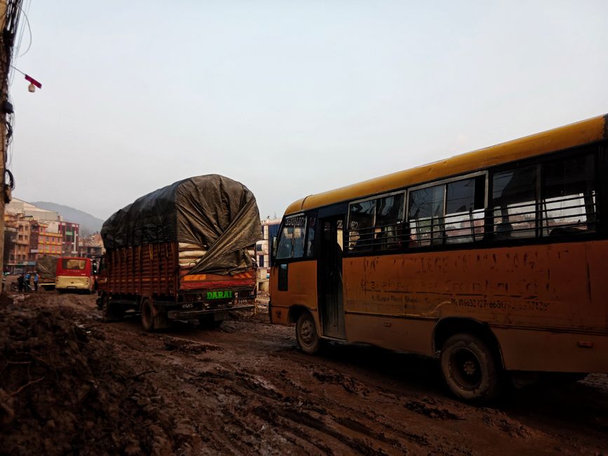 Buses and trucks navigate a muddy urban road, highlighting city travel challenges.