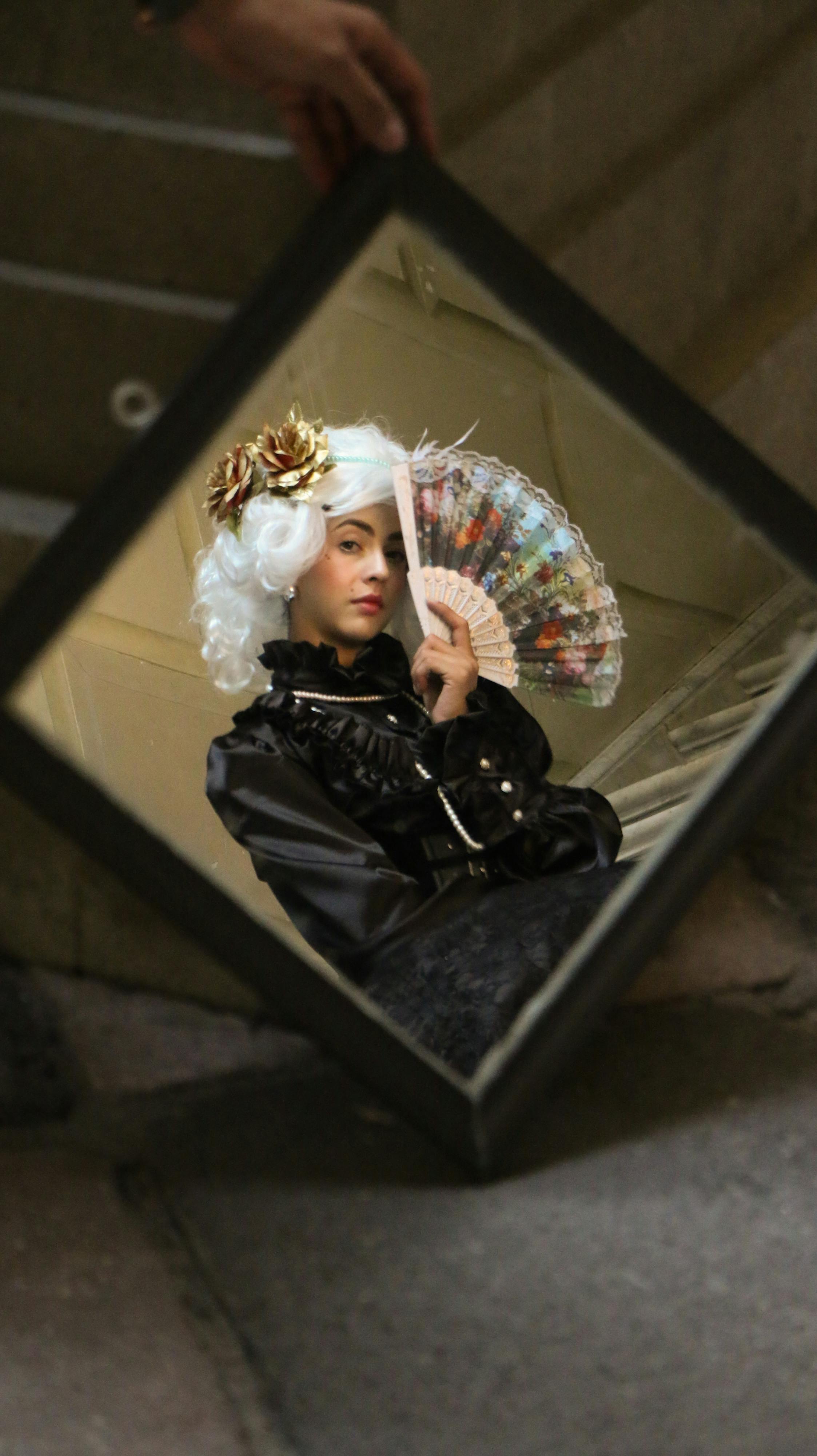 A woman in historical costume with a fan is reflected in a mirror, offering a whimsical, elegant scene.