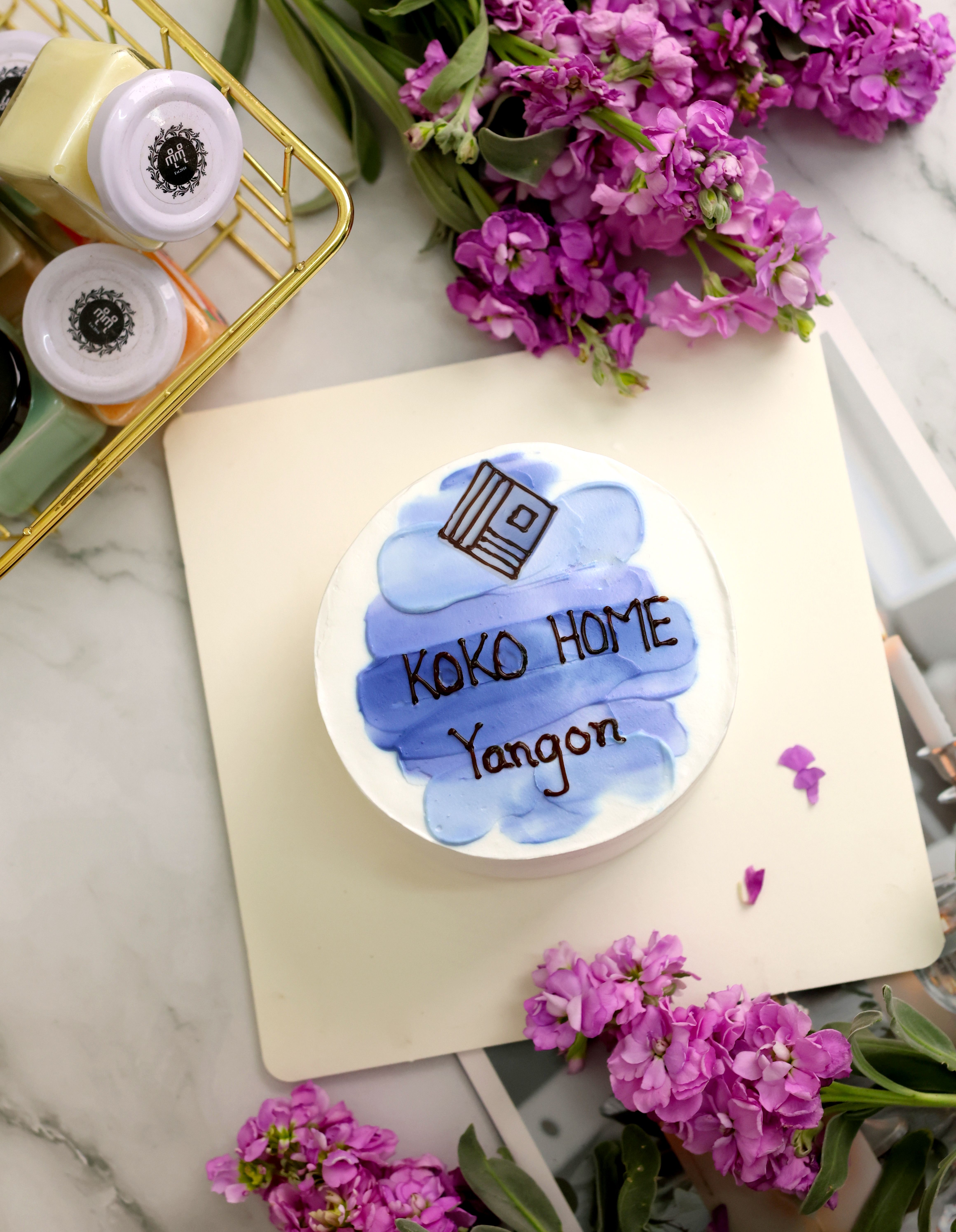 Elegant cake with KOKO HOME Yangon design surrounded by purple flowers on a marble surface.
