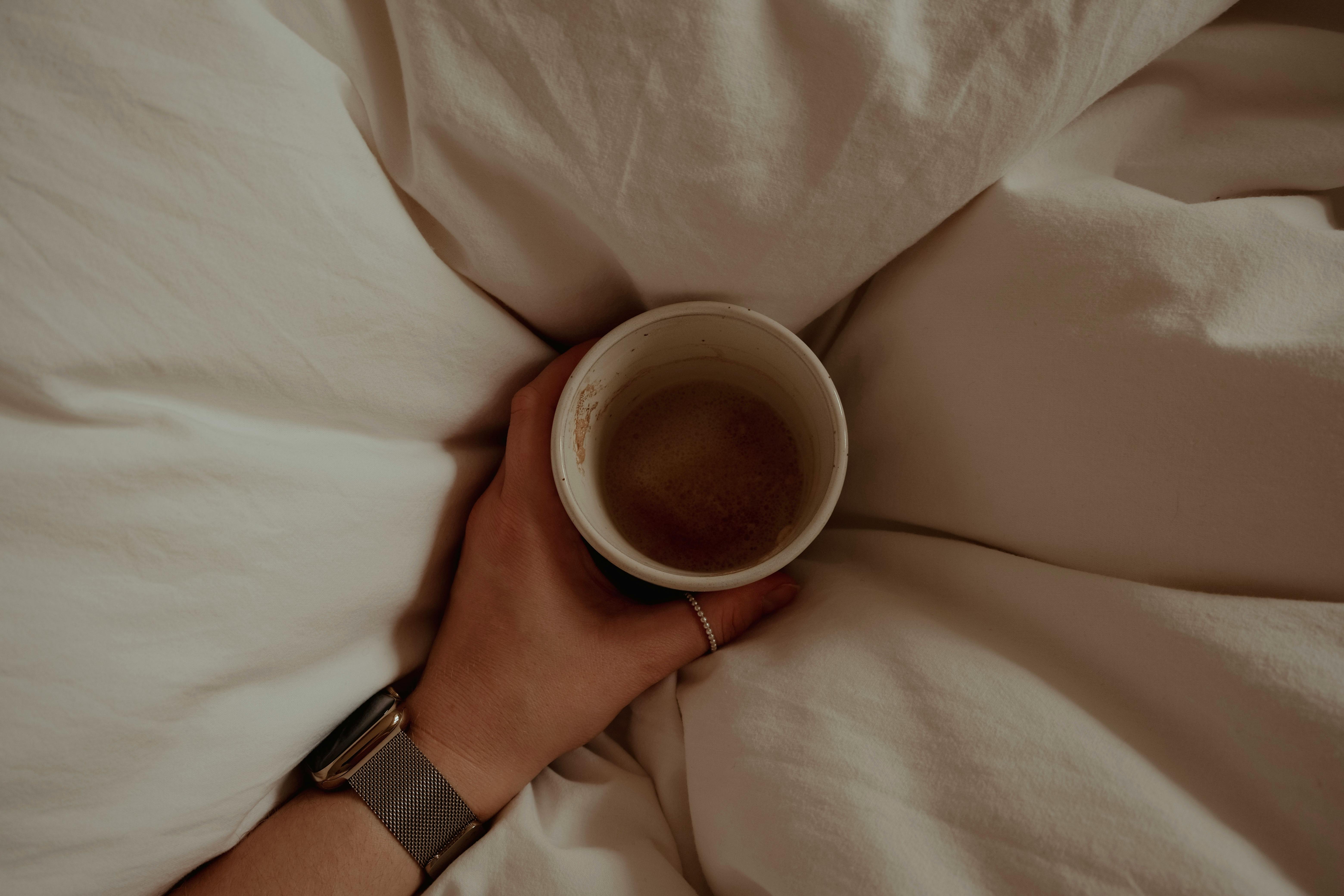 A relaxed morning scene with a person holding coffee in bed, surrounded by soft sheets.