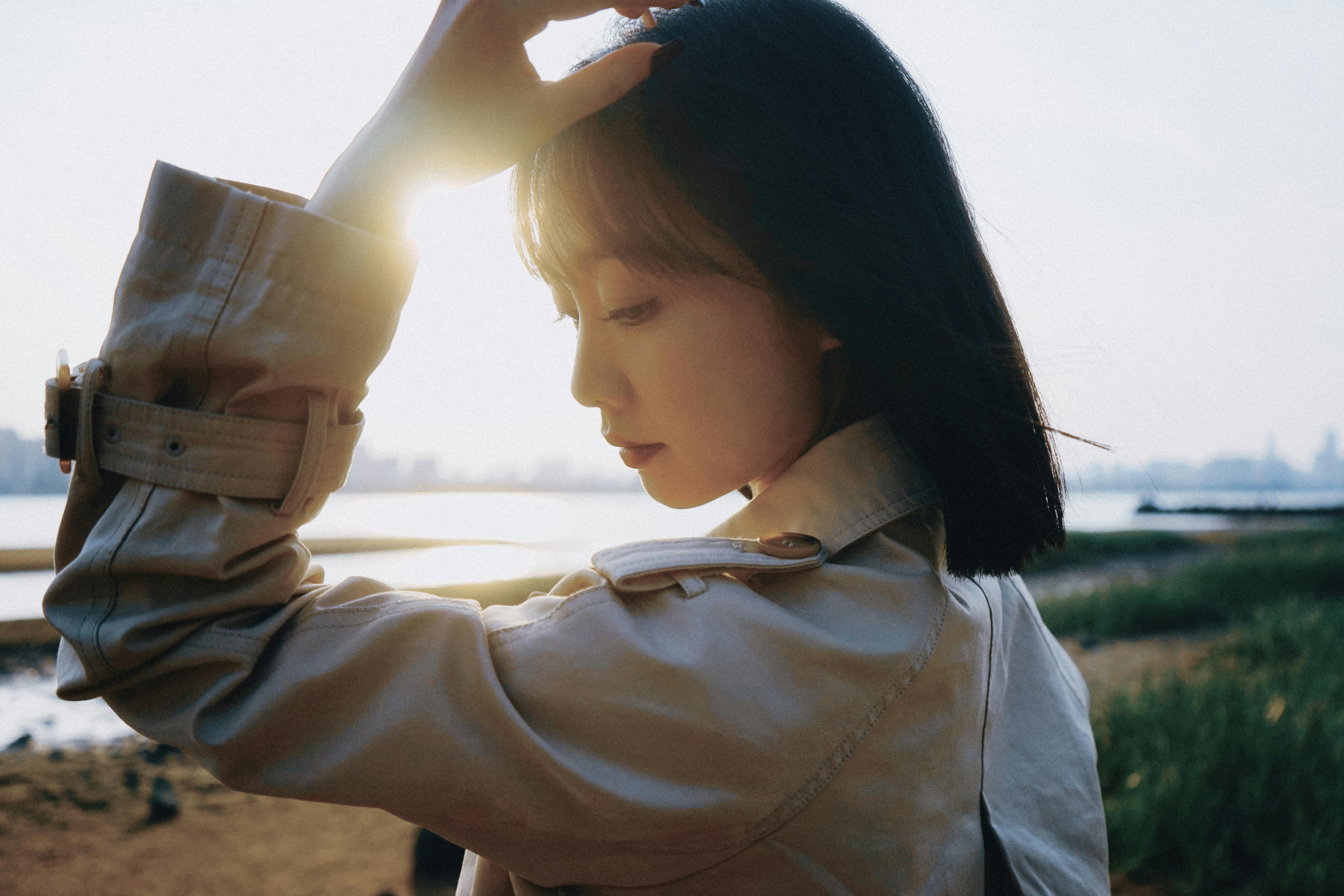 A beautiful woman in a beige coat by the beach with sunlight illuminating her face.