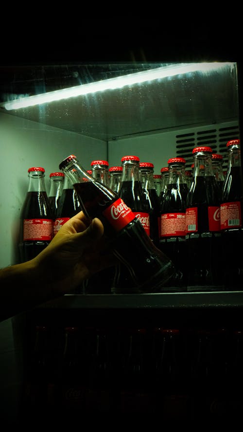 A person holding a bottle of coke in front of a refrigerator