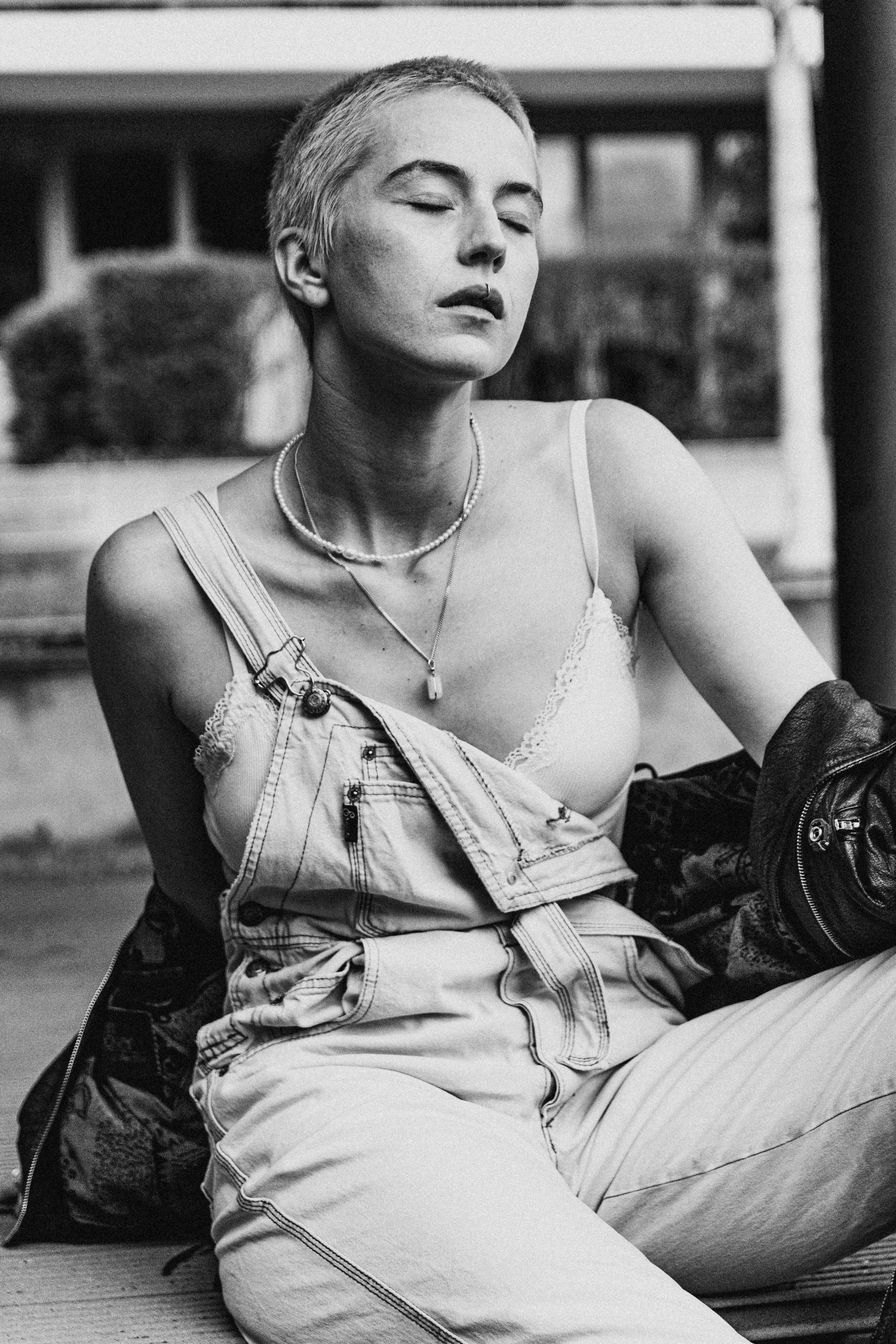 Free Black and white portrait of a woman wearing denim overalls and a necklace, posing outdoors. Stock Photo