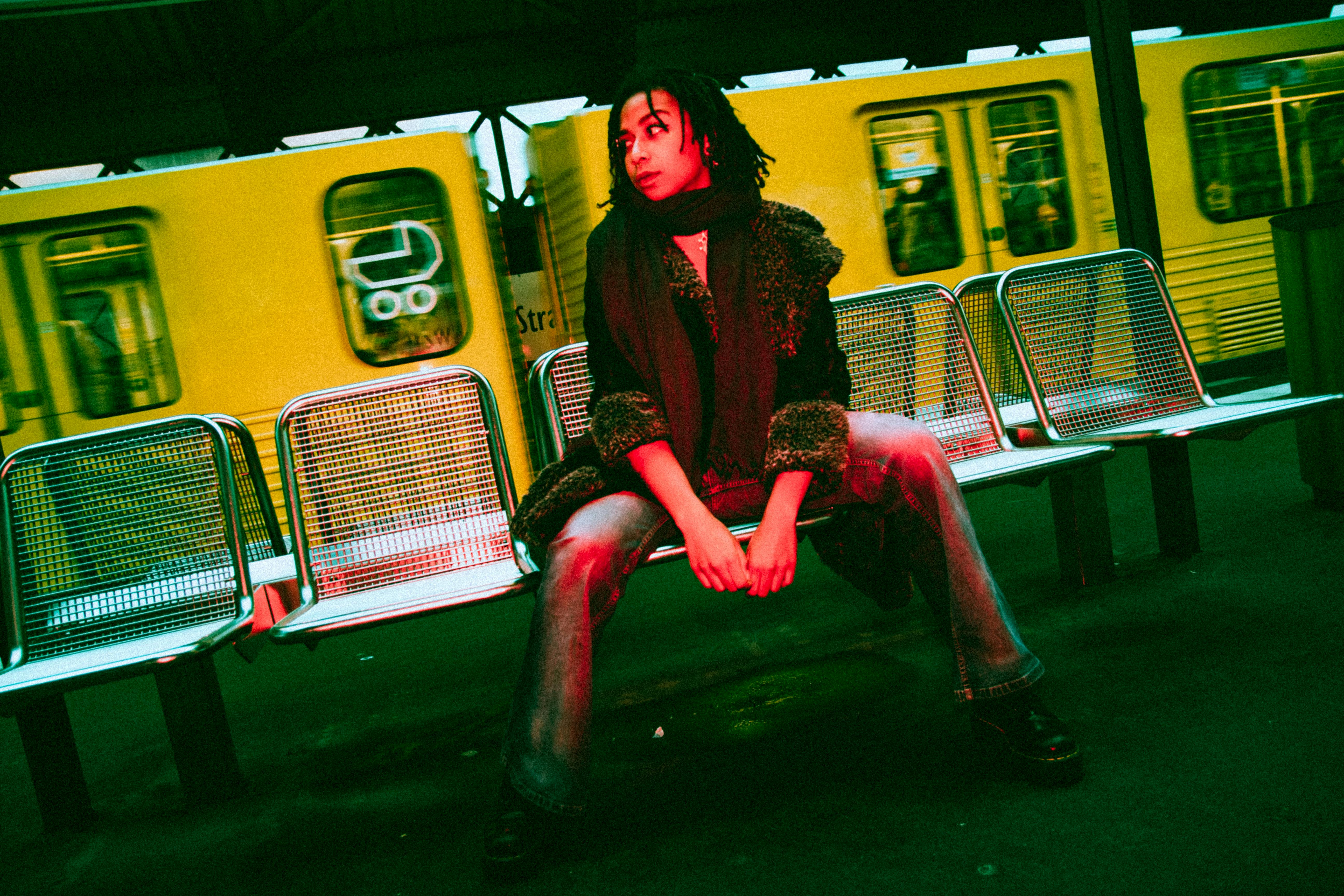 Stylish woman sitting on bench at Berlin train station with yellow train passing by, capturing urban fashion vibe.