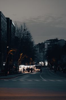 Moody evening street scene with people and trees silhouetted against city lights.