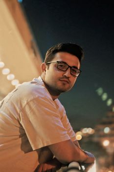 Portrait of a young man with glasses leaning on a railing in a city at night, surrounded by urban lights.