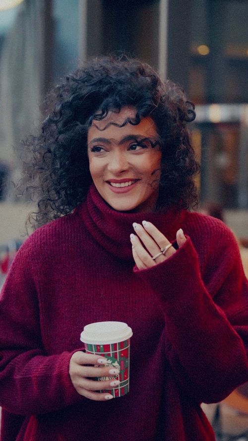 Free Cheerful woman with curly hair holding a festive cup, wearing a cozy sweater. Stock Photo