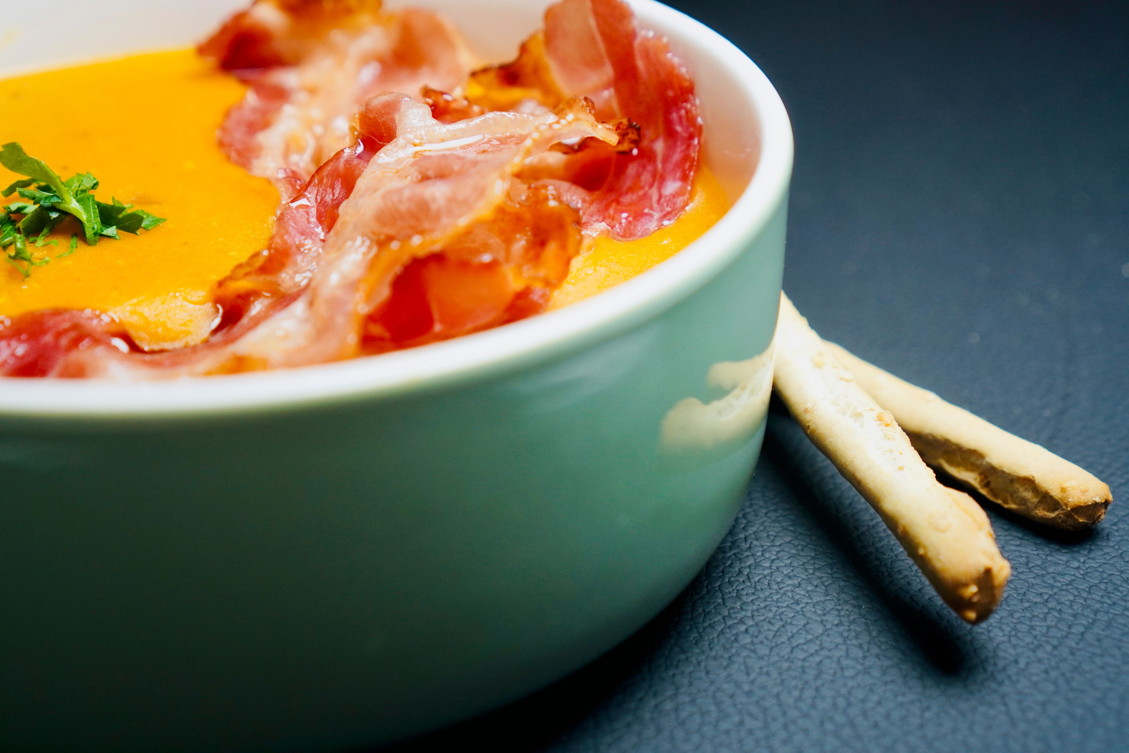 A close-up of a creamy orange soup topped with crispy bacon and herbs in a ceramic bowl (food photography).
