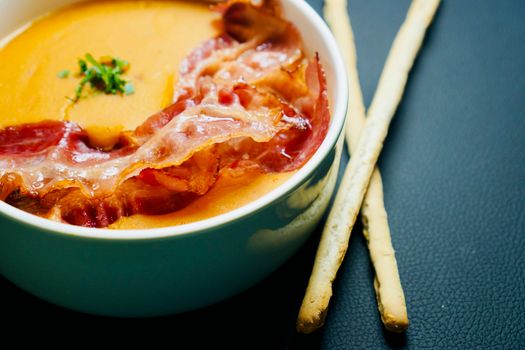 Delicious bowl of creamy soup topped with crispy bacon served with breadsticks on a dark background.