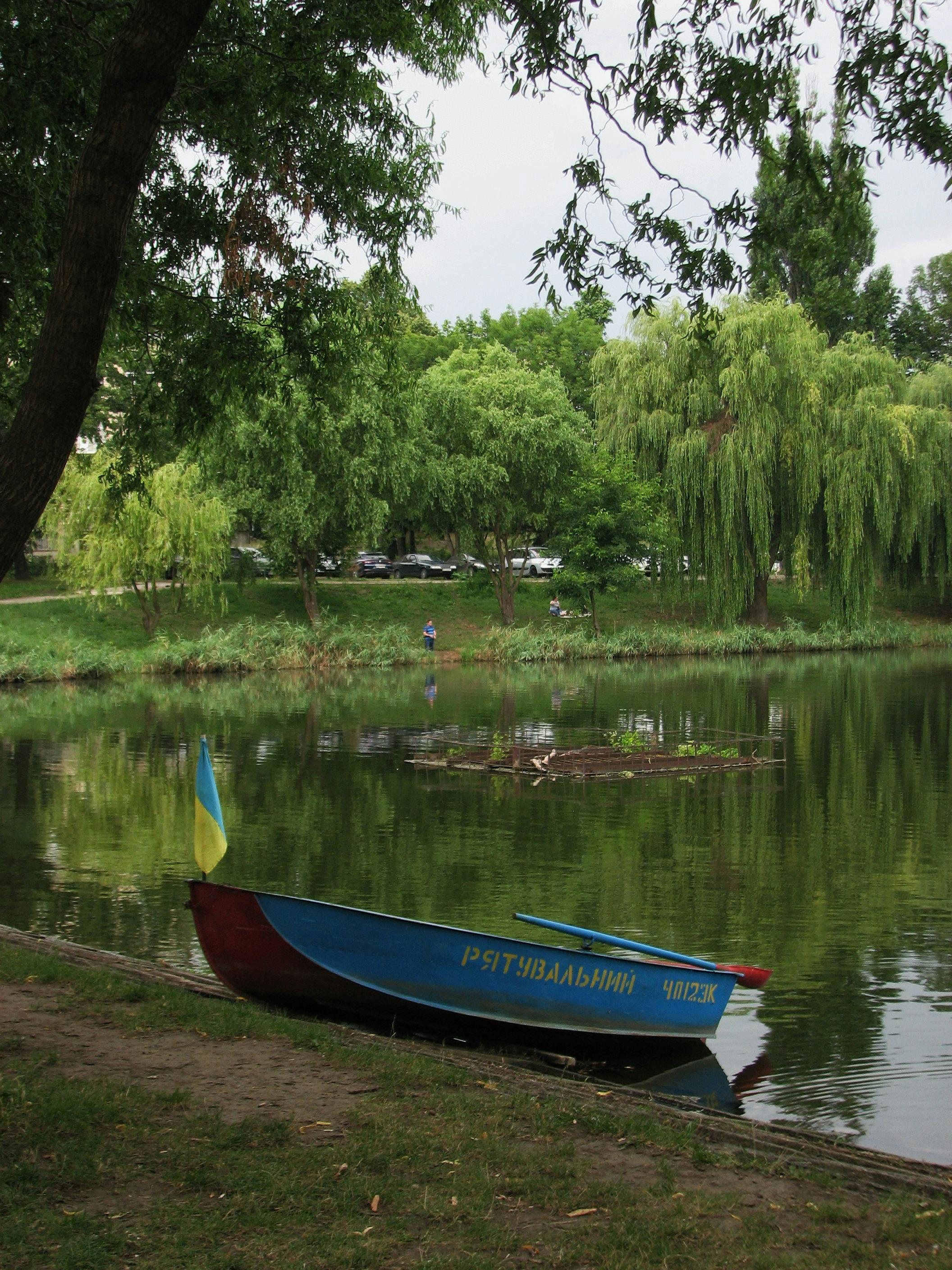 A tranquil scene of a boat by a pond with lush greenery in a park, featuring a Ukrainian flag.